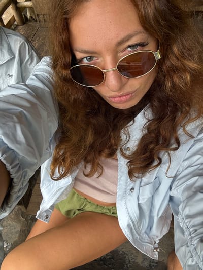 A woman with curly brown hair wearing sunglasses is posing for a picture. She is wearing a light blue, button-down shirt, a light pink top, and green shorts. The woman has fair skin and green eyes. The background consists of a weathered wooden surface and a textured, grey stone-like surface. The image is angled. The picture may have been taken in Yên Minh Commune, Vietnam.
