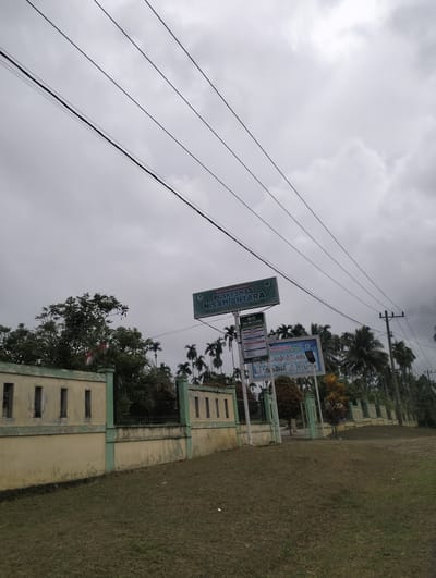 The image shows the entrance to a public health center, "Puskesmas Samantara," in Aluepapeun, Indonesia. A light yellow wall with green trim runs along the foreground, separating a grassy area from the facility. Several power lines are visible crisscrossing the overcast sky. Behind the wall, a gate leads to the Puskesmas. Two large billboards are displayed near the entrance, one with "Puskesmas Samantara" and another featuring information about healthcare services. Palm trees and other foliage are visible behind the building. The sky is heavily clouded, suggesting an overcast or potentially rainy day. No people are visible in the scene.