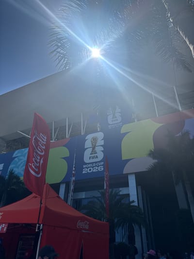 In Miami Gardens, Florida, a bright sun shines over a stadium decorated with a banner for the FIFA World Cup 2026. A red Coca-Cola flag and a red tent are in the foreground. The banner features the World Cup trophy and the number "26". Palm trees are visible throughout the scene.