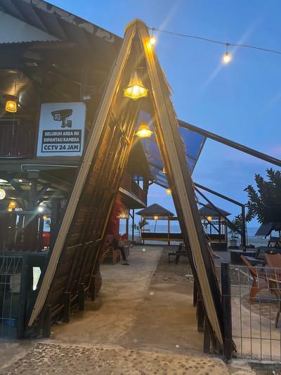 An outdoor seating area at dusk in Keudekruenggeukueh, Indonesia, features a prominent triangular entranceway made of wood and thatch. String lights with woven shades illuminate the pathway, and a larger caged light fixture hangs from the roof of the building on the left. A sign on the building states, in Indonesian, "SELURUH AREA INI DIPANTAU KAMERA CCTV 24 JAM" (This entire area is monitored by CCTV 24 hours). Beyond the entrance, the scene opens to a beach with two covered gazebos and tables and chairs set up on the sand. The sky is a deep blue with hints of pink and orange, indicating the setting sun. A person is partially visible sitting at a table on the right side of the image, and another person is seated on the left. The ground is a mix of concrete and small stones.