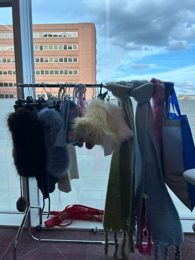 The image depicts an interior space overlooking an urban environment in Garston, Watford, United Kingdom, with a view of a multi-story building resembling structures found at the Building Research Establishment (BRE) campus. In the foreground, a metallic, wheeled clothing rack is positioned. This rack holds a variety of garments and accessories, including dark brown and grey faux fur pieces, knitted items in shades of light blue, white, and grey, and a prominent fluffy cream-colored item with an attached white tag. Multiple scarves in light green, light blue, and pink, some with braided fringes, are also present. Two shopping bags, one blue and one grey/silver, hang on the right end of the rack. Below the rack, on a dark red/purple carpet with a subtle geometric pattern, a red bag bearing the "MOSCHINO" logo is visible. Through the window, a large, rectangular, reddish-brown building with numerous horizontally aligned windows is seen under a sky with both blue patches and white clouds. No people are depicted.