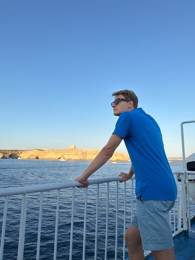 A young man with short blond hair and sunglasses stands on the deck of a ferry, leaning against a white railing. He is wearing a bright blue polo shirt and light grey shorts. The man is looking out towards a scenic coastline with rocky cliffs, a fort, and a few boats on the water. The sky above is clear and blue, and the water is a deep blue. The image captures a serene moment of travel, likely in the vicinity of Għajnsielem, Malta, given the distinctive coastal landscape.