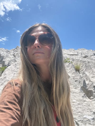 A woman with long blonde hair and sunglasses stands in front of a rocky, mountainous landscape under a blue sky with scattered clouds. She is wearing a brown t-shirt. The terrain behind her is rough and textured, with patches of green vegetation growing on the rock face. The image appears to be taken outdoors in a natural setting.