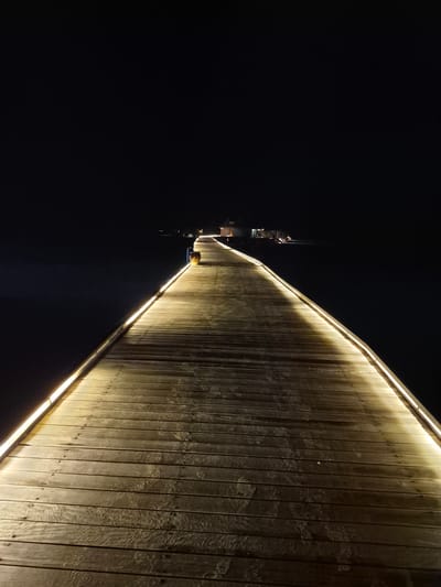 At night, the image depicts the illuminated boardwalk of the Bodufolhudhoo harbor in the Maldives. A wooden boardwalk runs horizontally across the frame, lit by strips of white-yellow lights. The left side of the frame is dark, suggesting an ocean or other body of water. A building, possibly a small structure with multiple levels, is visible in the distance to the left.