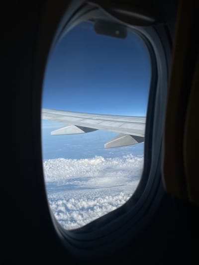 The image is an interior view from an airplane window. A portion of the airplane wing extends into the frame from the left. The wing is light gray and features a downward-angled flap near its end. Below the wing, a vast expanse of white, puffy clouds stretches across the scene. The sky above is a clear, deep blue. The window frame, which is dark and oval-shaped, partially obscures the view. The right side of the frame shows a glimpse of a seat, upholstered in a yellowish-brown fabric. The perspective suggests the airplane is in flight, high above the clouds. The location in the city of Nagyhajmás, Hungary is not discernible from this image.
