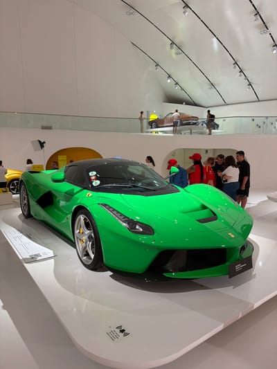 The image shows a bright green Ferrari LaFerrari on display at the Museo Enzo Ferrari in Modena, Italy. The car is parked on a white pedestal in a modern museum setting with a white, arched ceiling and exposed track lighting. In the background, on an upper level of the museum, another classic car, possibly a Ferrari, is visible, along with several visitors observing the exhibits. In the foreground, near the green Ferrari, a small group of people are gathered, some looking at the car and others talking. There are also informational plaques near the car, one of which reads "MUSEO ENZO FERRARI MODENA". Another plaque on the pedestal warns "Non toccare. Sine allarmi. Alarm activated."