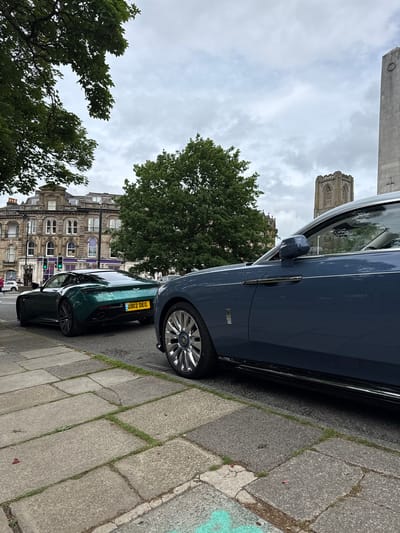 The image, captured in Harrogate, United Kingdom, depicts an urban street scene under an overcast sky. The original media file is rotated 90 degrees counter-clockwise.

When viewed upright, two luxury cars are prominently parked parallel to a stone-paved sidewalk on the right side of the street. In the foreground, a dark blue Rolls-Royce showcases its side profile, including a chrome door handle, side mirror, and a reflective multi-spoke wheel bearing the Rolls-Royce emblem. Its elegant design and emblem are clearly visible. Parked directly behind it is a dark green Aston Martin, seen from its rear-three-quarter angle, revealing its sleek body and complex alloy wheels. The text "ASTON MARTIN" is visible on its boot lid, along with a yellow UK license plate displaying "DB12 DEG".

The sidewalk, made of weathered grey flagstones with grass growing in the cracks, runs alongside the vehicles. In the background, an ornate, multi-story building with numerous arched windows and classical architectural details stands, on which a "NatWest" sign is visible, indicating a bank branch. Further in the background, a tall, historic stone tower with Gothic-style features, possibly part of a church or cathedral, rises against the cloudy sky. Lush green trees with dense foliage are scattered throughout the scene, contributing to a verdant urban landscape. The soft, diffused daylight suggests a cloudy day.