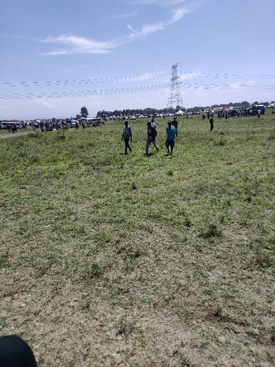 The scene depicts an open, sunlit grassy field in Olkaria ward, Kenya, beneath a clear blue sky with scattered white cirrus clouds. In the mid-ground, three adult males are observed walking from left to right, away from the camera's perspective. The leftmost individual wears a dark top and light trousers. The central figure wears a dark backpack and light-colored trousers. The rightmost individual wears a blue patterned shirt and dark trousers. Approximately 150-200 meters into the background, a large metal lattice electrical transmission tower stands prominently to the right of center, with power lines extending horizontally across the frame. Adjacent to and behind the tower, a dense assembly of approximately 50-70 parked vehicles, predominantly light-colored cars and vans, are visible along a treeline. Numerous individuals are interspersed among these vehicles, suggesting a gathering. A single white canopy tent is situated near the electrical tower within the vehicle area. The foreground consists of short green and brown grass with visible patches of dry soil. A dark, out-of-focus object occupies the bottom-left corner of the frame.