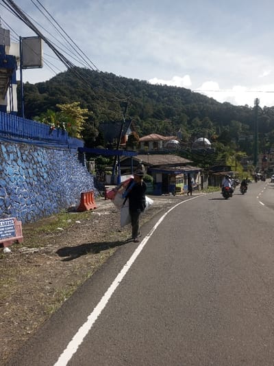 The image depicts a daytime street scene in a mountainous area, specifically identified as Ka Thaung (Upper), Myanmar. A paved, two-lane road curves slightly to the right, occupying the center and right portions of the frame. A continuous white line delineates the left edge of the roadway.
On the left side of the road, a dirt and sparse grassy verge extends towards a robust blue-painted stone wall with an irregular pattern, topped by a matching blue metal fence. An orange plastic traffic barrier is positioned on this verge, adjacent to the wall. A white rectangular sign with red text, partially visible and indicating a prohibition, is staked into the ground near the wall.
A lone individual, dressed in dark attire and wearing a hat, walks away from the viewer on the left side of the road, just to the right of the white line. This person is carrying multiple large, light-colored bags and other objects, some slung over their left shoulder. A long shadow cast by this individual stretches across the road and verge to the left, indicating a low sun angle.
Further down the road on the right, several motorcycles are visible, some with riders, moving away from the camera. Various buildings line the right side of the road, appearing to be commercial or residential structures, with some featuring multiple stories and a distinct domed roof visible on one.
The background is dominated by a large, densely forested mountain range rising steeply. Overhead, numerous black electrical wires crisscross between poles, originating from a structure on the far left. The sky above is partly cloudy with patches of blue visible.