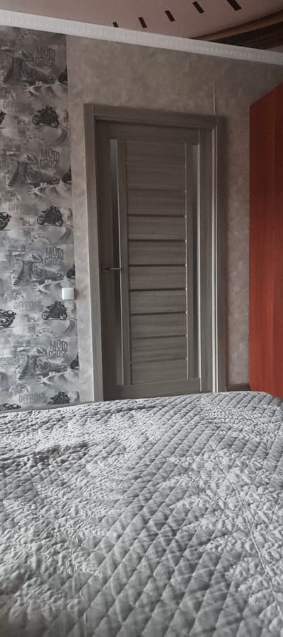 A slightly low-angle, eye-level shot captures a bedroom interior. On the left wall, patterned wallpaper depicts motorcycles and the words "MOTO CROSS" and "OFFROAD" in a grayscale, graphic style. A white square light switch is visible on the wallpaper. A light brown, modern interior door with horizontal slats is positioned centrally. To the right of the door, a portion of an orange wall is visible. In the foreground, a bed with a textured, quilted gray blanket fills the lower half of the frame. The scene appears to be staged and likely within a residential setting in Aktanyshbash, Russia.