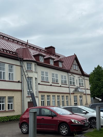 This image presents a multi-story building in Lappeenranta, Finland, under an overcast sky. The building exhibits a light yellow and cream-colored stucco facade with dark red trim detailing. Its most prominent feature is a complex, dark red standing-seam metal roof system, which includes multiple dormer windows with white frames and decorative cross-bracing, a rounded turretted section on the left, and a distinct gable end on the right featuring half-timbering.
A long, black, angled metal structure, resembling a roof hoist or a substantial extension ladder, is positioned against the left side of the building, extending from near ground level up to the red metal roof.
In the paved foreground, a red Peugeot 307 hatchback is parked, oriented towards the right, displaying Finnish license plate AZR-589. Directly behind it, a white hatchback is parked, followed by a partially visible dark SUV or van. A grey metal bollard or post is situated in the immediate foreground, partially obstructing the front left portion of the red car. Low green foliage is visible along the base of the building on the far left. A large, verdant deciduous tree stands on the far right of the frame. A single small, dark-colored bird is visible in flight in the upper central portion of the grey sky. No people are discernible in the scene.