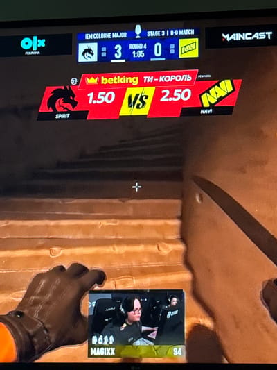 The image shows a screen displaying a live esports match, likely Counter-Strike: Global Offensive, given the logos and terminology. The match is identified as "IEM Cologne Major" and is in "Stage 3". The current score is 3-0, and the round timer shows 1:05. Two teams, "SPIRIT" and "NAVI", are competing, with "SPIRIT" having odds of 1.50 and "NAVI" having odds of 2.50. The screen also features logos for "OLX" and "MAINCAST", suggesting broadcasting partners or sponsors. In the foreground, a gloved hand is visible, along with a smaller inset screen showing two players, identified by the name "MAGIXX" and the number "94" displayed below. The background of the main screen appears to be a stylized, textured environment, possibly from the game itself. The location of Gothenburg, Sweden, is not depicted or suggested by the visual information in the image.