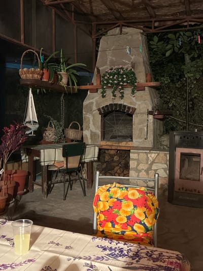 This is an exterior night shot of an outdoor patio area, likely in Kichevo, Bulgaria. The scene features a rustic stone oven and grill, a wooden table with chairs, and a covered seating area. The oven is central to the image, with a large fan in its opening and stacked firewood below. To the left of the oven, a wooden shelf holds potted plants and baskets, with a small model sailboat hanging nearby. A table covered with a checkered cloth is set with chairs, one of which is adorned with a colorful floral cushion. In the foreground, a table is partially visible, covered with a tablecloth featuring a lavender pattern, and a plastic cup filled with a light-colored liquid sits on it. The surrounding environment is dimly lit, with shadows cast by the overhead cover and surrounding foliage.