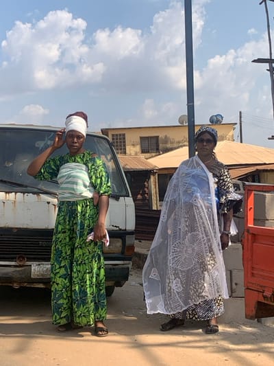 Two women stand on a street next to a dilapidated white van. The first woman wears a green floral dress with a white head wrap and sash. She is positioned in front of the van, holding a pink item. The second woman, located further back, wears a patterned blue dress, a matching head covering, and sunglasses. A white lace shawl drapes over her shoulders. The background features buildings under a partly cloudy sky. A utility pole stands to the left.