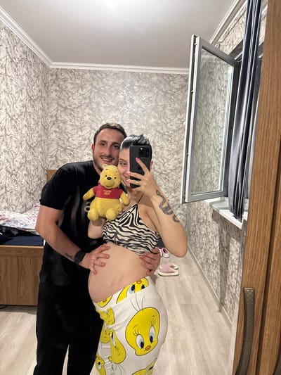 A pregnant woman and a man are posing for a mirror selfie in what appears to be a bedroom. The woman is wearing a black and white zebra-print top and yellow pants with a Tweety Bird pattern. She is holding a Winnie the Pooh plush toy. The man is wearing a black t-shirt and is standing behind the woman, with his arms around her. The room has textured wallpaper, a wooden floor, and a large mirror that reflects the scene. A window with grey curtains is visible in the background. A wooden dresser or bed frame is also present. There are pink sandals on the floor. The image captures a moment of anticipation and joy, likely in a domestic setting.