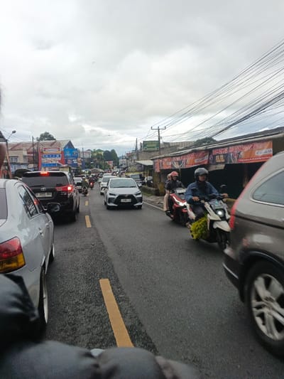 This image depicts a congested urban street scene in the city of Masaiti, Zambia, under an overcast sky. The view is from a low perspective, likely from a vehicle or on foot, looking down a two-lane road with a double yellow line marking the center.
In the immediate foreground, dark, possibly fabric-covered, elements partially obstruct the view at the bottom. To the left, the rear section of a silver sedan is visible. To the right, the front-right side of a dark grey SUV or minivan is partially captured, with its wheel in motion.
The midground features dense traffic. A black SUV with illuminated red taillights is positioned on the left lane, behind the silver sedan. Further down the left lane, several cars and motorcycles are either stopped or moving slowly. In the right lane, two motorcycles are prominent: the foremost, a white scooter with a male rider in a blue jacket and dark helmet, transports green leafy items attached to its side. Behind him, a red scooter carries a driver and a female passenger wearing a pink helmet and light-colored clothing. A white sedan is visible in the distance, further contributing to the traffic.
The background comprises a street lined with buildings. On the left, multi-story structures display various commercial signs, including "Indomaret" and "MASPION". On the right, lower-rise buildings feature banners, one clearly reading "BENGKEL MOTOR AHASS" (Motorcycle Workshop) and mentioning "Jalan Raya Parakan Tasikmalaya". Numerous overhead power lines and cables crisscross above the buildings. The sky is uniformly grey and cloudy. Distant hills or mountains are faintly visible beyond the urban sprawl.