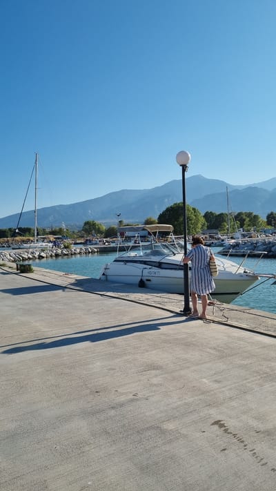 The image depicts a scenic view of Limani Litochoro, Greece. In the foreground, a concrete pier runs along the water's edge, casting long shadows from a tall lamppost. A woman, wearing a blue and white striped dress and carrying a woven bag, stands near the lamppost, looking down at a white motorboat docked beside the pier. The boat, identified by the registration "HD-TW 71," is the prominent subject in the middle ground. In the background, a calm body of water reflects the clear blue sky. Further back, a sailboat with its mast prominently visible, a rocky breakwater, and a line of trees suggest the edge of the harbor. The distant landscape is dominated by a range of majestic mountains under a cloudless sky, indicative of the natural beauty surrounding Litochoro. Several other boats are visible moored in the harbor, adding to the maritime atmosphere of the location.
