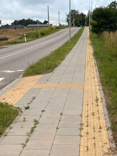 The image depicts a sidewalk alongside a road in Aukštieji Rusokai, Lithuania. The sidewalk is paved with light gray square tiles and features a tactile paving strip in a yellow color, designed for visually impaired pedestrians. The yellow strip runs along the edge of the sidewalk closest to the road. The road itself is paved and has white dashed lines indicating lanes. To the left of the road, there is a grassy verge and further left, a construction site with some buildings under construction and a white road marker. To the right of the sidewalk, there is a more substantial grassy area with tall grass and some trees in the distance. The sky is overcast. There are no people visible in the image. The scene is an outdoor urban environment with signs of infrastructure and development.