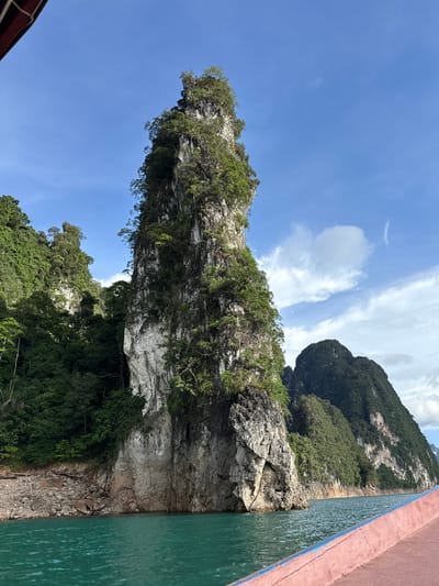 The image shows a rocky, jungle-covered karst formation rising out of green, calm water. The rock face is steep and weathered, with dense vegetation clinging to its crevices. The sky is a clear, bright blue with a few wispy white clouds. The perspective suggests the viewer is on a boat, looking towards the shore. To the right of the frame, the side of a boat with a reddish-brown stripe and a green hull is visible, with ripples on the water indicating movement. The overall scene is one of natural beauty and tranquility, possibly a tourist or scenic location. The lighting and bright sky suggest it is daytime, likely mid-morning or afternoon. The lush greenery hints at a tropical climate. The location is identified as Khao Phang, Thailand. No people or text are visible.