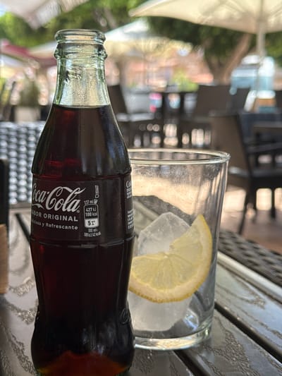 A close-up, rotated shot shows a glass bottle of Coca-Cola Original Flavor and a nearly empty glass of soda with ice and a lemon slice. The Coca-Cola bottle is dark brown and features a black and white label with the iconic Coca-Cola logo, along with nutritional information in Spanish. The label states "SABOR ORIGINAL" and "Deliciosa y Refrescante." It also indicates "237 ml," "427 kJ / 100 kcal," and "5%" for a serving of 100 ml. The glass is clear and contains ice cubes and a thin slice of lemon. The scene appears to be outdoors, possibly at a cafe or restaurant, with a blurry background suggesting trees and outdoor furniture. The lighting suggests it's daytime. The overall mood is casual and relaxed. The text "Campillo Del Moro, Spain" is a caption and not visible in the image.
