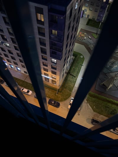The image is an aerial view of a residential complex at night, likely in Perm, Russia, as indicated by the prompt. The scene captures the side of a modern apartment building with numerous illuminated windows. Below the building, a paved area with parked cars is visible, along with a section of green lawn and a children's playground. The foreground is partially obscured by dark blue metal railings, suggesting the photo was taken from a balcony or window. The lighting suggests it is evening or night, with artificial lights illuminating the buildings and playground area.