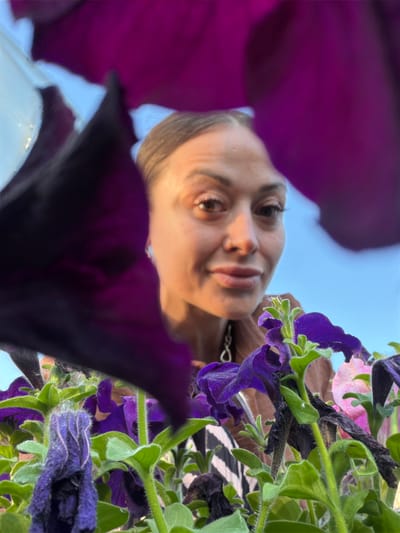 A close-up, low-angle shot captures a woman from the chest up, peering through a dense cluster of purple petunias. Her face, softly lit by warm, possibly late afternoon sun, is turned towards the camera. She has light brown hair pulled back, prominent eyebrows, and her eyes are directed at the lens. Her lips are slightly parted in a gentle smile. She is wearing a brown top and a necklace with a circular pendant. The foreground is dominated by the vibrant purple and green foliage of the petunias, with some flowers appearing wilted or in bloom. The background is a soft blur of light blue sky. The overall impression is an intimate, perhaps candid, moment shared amongst flowers.