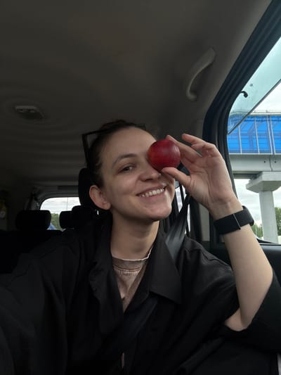 A woman is inside a vehicle, holding a red nectarine to her nose as if to resemble a clown's nose. She is wearing a black watch and a black overshirt and seatbelt. The interior of the vehicle is predominantly gray. Through the car window, a blue glass structure is visible, potentially located in Baskan, Russia.