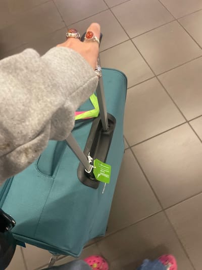 The image displays a downward view of an unidentified individual's right arm and hand, which is gripping the extended, grey two-pole handle of a teal-colored soft-sided rolling suitcase. The person is wearing a light grey long-sleeved garment, with the sleeve covering the forearm. Three rings are visible on the right hand: one large reddish-orange stone ring on the index finger, a smaller reddish-orange stone ring on the middle finger, and a ring with multiple clear stones on the ring finger.
Attached to the black handle mechanism of the suitcase is a bright green luggage tag displaying the "Ethiopian" Airlines logo. A neon yellow-green strap or piece of fabric is also visible behind the suitcase handle. At the bottom of the frame, parts of the individual's lower legs and feet are visible, wearing blue jeans and pink Crocs-style clogs. The environment consists of a light grey tiled floor with darker grout lines. This scene is consistent with an airport terminal, specifically O.R. Tambo International Airport in Kempton Park, South Africa, given the presence of an "Ethiopian" Airlines tag.