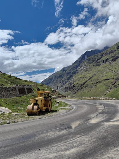 This is a landscape photograph taken in Yari-Khoksar, India. A bright yellow road roller is parked on the side of a paved, winding mountain road. The road roller is a large piece of construction equipment with a heavy cylindrical drum in the front and wheels in the back. The road itself appears to be under construction or repair, with patches of loose gravel and sand on the asphalt.

To the left of the road, a steep, grassy embankment rises, supporting a low stone wall with concrete reinforcements. Beyond this, more green, grassy hills stretch upwards.

To the right of the road, a massive, steep mountain face dominates the scene. Its slopes are covered in green vegetation, with rocky outcrops and crevices visible. The mountain rises sharply into a cloudy sky. The sky is a vibrant blue, dotted with large, fluffy white clouds that cast shadows on the mountain. The overall impression is of a remote, high-altitude location, possibly undergoing road maintenance.