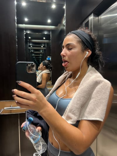 The image captures a young adult female inside an elevator in Bağcılar, Turkey. The subject is depicted from the chest up, in profile facing right, with her tongue extended. She exhibits visible perspiration on her forehead, temples, neck, and décolletage. Her dark hair is pulled back and held by a black headband. She is dressed in a light blue sports bra and gray athletic bottoms, with a light tan towel draped over her shoulders. White wired earphones are inserted into her ears. In her left hand, she holds a dark-colored smartphone with a prominent multi-lens camera array, positioned as if for a selfie, and a partially full clear plastic water bottle with a blue cap. Her fingernails are painted with a dark, patterned polish. The elevator interior features dark, highly reflective surfaces, including mirrors that produce an infinite reflection effect, and recessed overhead lighting. A metallic elevator door panel is visible on the right side of the frame.