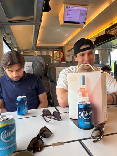 Two young men are seated on a train, likely in Norway. They are positioned at a table between them, which holds two blue cans of Hansa beer, a box of Ioppa Rosato wine, and a pair of sunglasses. One man, wearing a black baseball cap and a white t-shirt, is looking towards the camera with a slight smile. He has tattoos on his arm. The second man, wearing a navy blue polo shirt, appears to be sleeping or resting with his eyes closed. He has brown hair. The train interior is visible, with seats and overhead compartments. A digital screen on the wall displays train route information, with stations listed including Moss, Ryggre, Radie, Fredrikstad, and Halden. The lighting suggests it is daytime. The overall atmosphere appears relaxed.