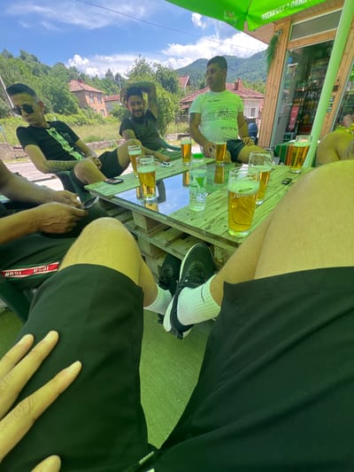 The image depicts an outdoor social gathering of at least five adult males in Касапите, Bulgaria, under a bright, sunny sky. The scene is set on a green ground surface, possibly artificial turf, around a rectangular table made of light-colored wooden pallets with a glass top. A large green patio umbrella, bearing upside-down Cyrillic text, provides shade over the table.

To the right of the frame, a single-story commercial building with a wooden facade and large glass display windows is visible, featuring Cyrillic text (e.g., "КЛИМБО 24 ЧАСА" and a Coca-Cola logo inside). The background reveals a rural or semi-rural landscape with rolling green hills, dense trees, scattered white cumulus clouds in a blue sky, and several traditional houses with red tile roofs.

Five men are seated on green chairs around the table. In the immediate foreground, the legs of two individuals are visible: one on the right wears black shorts, white Nike socks, and black sneakers; another on the left wears black shorts with a red and white "ICON" stripe and has a left hand resting near a mobile phone.

Moving towards the mid-ground, a young male on the left wears black sunglasses, a black graphic t-shirt, and black shorts, displaying arm tattoos; he leans back, looking downwards. Next to him, a male with dark hair and beard, wearing a dark t-shirt and shorts, exhibits extensive arm tattoos and smiles slightly while looking towards the viewer, with his left arm raised behind his head. To the right, another male with short dark hair wears a white t-shirt with black text ("THE GHOST...WITH THE MOOD") and dark shorts, showing a left arm tattoo; he sits upright, looking left. Portions of a sixth individual are also visible on the far right.

On the table are six glasses of light amber liquid with white foam, consistent with beer, one of which clearly displays "ПИВО Лагер" in Cyrillic. A 500ml plastic water bottle with a green cap, two smartphones (one black, one silver), a red cigarette lighter, a white cigarette lighter, and a set of keys are also present on the tabletop. The individuals are in various relaxed postures, with some observing their surroundings and others interacting with their phones.