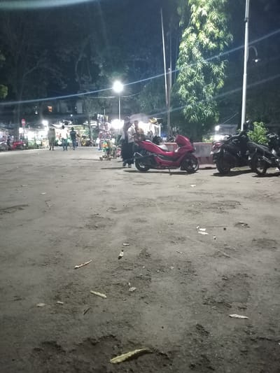 The image portrays a nighttime scene in a busy public space within the city of Nganjuk, Indonesia. The immediate foreground is an unpaved or heavily worn asphalt surface, dark grey in color, characterized by numerous visible potholes, cracks, and scattered debris including dried leaves. In the midground, a red scooter, identifiable as a Honda PCX or similar model, is parked facing left. To its right, two black motorcycles are parked, one facing right and the other partially obscured. A low, light-colored concrete barrier runs behind these vehicles.
Several individuals are present, indicating active foot traffic and social gathering. Towards the right midground, a group of people is gathered near an illuminated vendor stall; one woman, wearing a light top and a hijab, is clearly visible facing right, potentially interacting with the vendor or other individuals. Further to the left and deeper into the scene, multiple other figures are seen walking away from the camera.
The background is dominated by a line of brightly lit vendor stalls, indicating a night market or a street food area, with various goods displayed. Tall trees with dense foliage are visible on both sides of the frame, partially silhouetted against the artificial light sources. Multiple streetlights and individual stall lights illuminate the scene, casting a warm, yellowish glow and creating visible lens flares. A prominent tall utility pole with an attached lamp is visible on the right, and another on the left. A circular white and red sign is partially visible on the far left. The overall environment suggests a bustling nocturnal commercial and social hub.