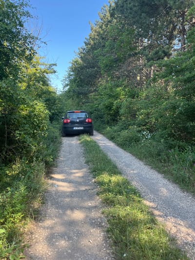 A black Volkswagen Golf is parked on a dirt road surrounded by lush green trees and foliage. The car has a license plate that reads "A 5114 PM". The road appears to be a path through a forest or wooded area. The sky is clear and blue. The scene is consistent with an outdoor location, possibly in a rural or natural setting near Kichevo, Bulgaria.