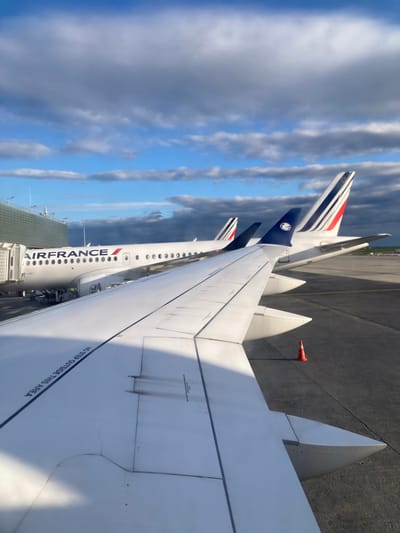 The photograph shows a view from an airplane wing, likely taken at Charles de Gaulle Airport (CDG) in Le Mesnil-Amelot, France. Two Air France planes are visible, their fuselages bearing the airline's name and logo. The tail fins are painted with the French tricolor. The focus is on the wing of the aircraft from which the picture was taken, displaying a "No Step Outside This Area" marking. The sky is filled with clouds, and a section of airport tarmac can be seen alongside a small orange cone. The buildings surrounding the airport are visible in the background.