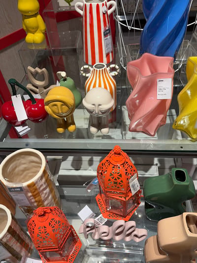 This media file is a close-up, eye-level shot of a retail display of decorative items, likely in a store in Pekalongan, Indonesia. The display features a variety of ceramic and glass objects arranged on shelves, with some items placed on clear acrylic risers.

Prominently visible are several colorful vases and decorative pieces. On the left, a large, twisted blue glass vase is partially visible at the top, followed by a soft pink, star-shaped glass vase with a price tag indicating "informa" and "Glass Vase Twis Table Top Decor Rp 100.000". Below these, a striped ceramic vase with red and white horizontal stripes is displayed alongside another ceramic item with orange and white stripes.

To the right, a bright orange, ornate lantern with intricate cut-out patterns is a focal point. Next to it, a set of pale pink, wavy, abstract decorative objects are arranged. Further to the right, a green ceramic vase with a unique, sculptural form is seen, as well as a tan ceramic planter.

At the bottom of the frame, a yellow, stylized figure with large, round limbs is displayed, and below that, a pair of bright red ceramic cherries with a green stem and a white tag. A large, beige ceramic vase with light brown stripes is also visible in the lower right. The overall impression is of a vibrant and varied collection of home decor items. The lighting suggests an indoor setting, likely a well-lit retail environment. There are no people visible in the image. The visible text includes brand names and product descriptions.