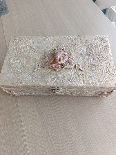 A vintage, ornate, rectangular wooden box with a textured, decorative surface in a pale gold and cream color scheme sits on a light brown wooden table. The box has a hinged lid with a metal clasp in the center. The lid is adorned with a raised, decorative design featuring an elephant-like motif in a pinkish-brown color, surrounded by an intricate, embossed pattern of swirling paisley and floral elements. The box appears to be a decorative storage container. The environment suggests an indoor setting, possibly a home. There is no identifiable location in the city of Toruń, Poland, or any people or actions present in the image.