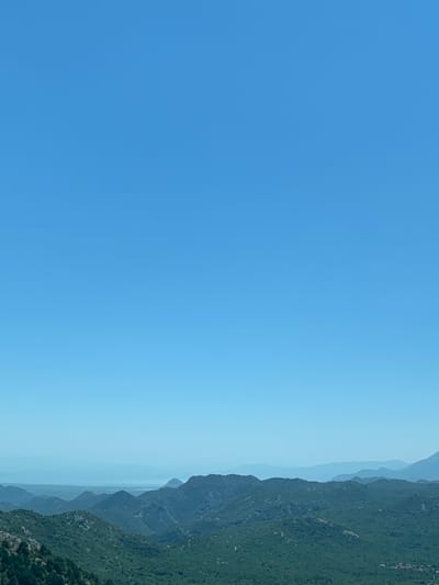 The image depicts a panoramic view of a mountainous landscape with a clear, bright blue sky dominating the upper portion. The foreground features lush green, densely vegetated hills with rugged terrain. In the mid-ground, a series of rolling hills and mountains recede into the distance, their surfaces covered in similar vegetation. Further in the background, layers of hazy, blue mountains are visible, creating a sense of depth. The furthest horizon suggests a body of water or a vast expanse, also rendered in shades of blue and fading into the atmospheric haze. No people, structures, or specific objects are discernible within the scene, focusing solely on the natural environment. The scene is likely captured from an elevated viewpoint, offering a sweeping vista of the natural topography. The location is identified as Dobrsko selo, Montenegro.