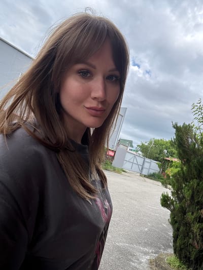 A photograph shows a woman with brown hair and green eyes, positioned in the foreground with a slight tilt. She wears a black shirt. Behind her is an outdoor scene in what appears to be the city of Khadyzhensk, Russia, indicated by signs in Russian. A grey road extends into the background, with a white fence and buildings visible along the side. Trees and greenery are present. The sky is overcast.