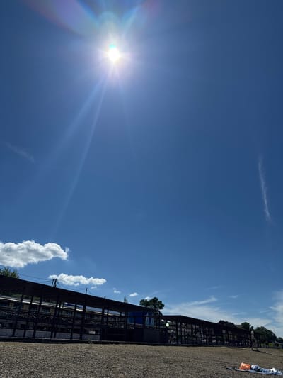 The image, rotated 90 degrees clockwise, depicts a sunny outdoor scene under a clear blue sky with scattered white cumulus and wispy cirrus clouds. The sun, prominently positioned in the upper left, generates a strong lens flare and visible rays.

A long, dark, open-sided structure, possibly a shaded walkway, stable, or market stall, extends horizontally across the middle ground. This structure features a slatted or beamed roof supported by dark vertical frames, and it casts shadows. Several people are discernible within and near the structure, with one individual standing near its far end. The foreground is covered with gravel. In the far right section of the gravel, an orange item (possibly a bag or tarp) and a white/blue item are resting. Green trees and foliage are visible beyond the structure and in the background.

The setting appears to be an outdoor public or recreational area, likely in Taman, Russia, during a bright day. No discernible text is visible in the image.
