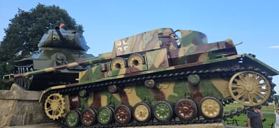 This is a medium-shot, eye-level photograph taken outdoors, likely during the daytime, featuring two historical tanks positioned on a stone foundation under a clear blue sky. The tank in the foreground is painted in a camouflage pattern of green, brown, and tan, and appears to be a German Panzer. It has a series of road wheels and tracks visible. A prominent white Iron Cross insignia is visible on its side. The tank in the background is painted a solid dark green and exhibits a red star insignia on its turret, indicating it is likely a Soviet T-34 tank. The tanks are situated in what appears to be an outdoor memorial or museum area. In the lower right corner of the frame, two individuals are partially visible; one man wearing a patterned shirt and sunglasses is looking towards the tank, and a woman in a light-colored top is standing next to him. A large tree with green foliage is visible behind the Soviet tank on the left. This scene is located in the city of Svidník, Slovakia, which is known for its significant Soviet military history and memorial sites.
