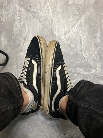 The image displays a pair of black and white Vans Old Skool sneakers worn by an individual whose lower legs and feet are visible. The shoes are positioned side-by-side, with the laces tied. The right shoe shows more wear and dirt on its white sole and side stripe compared to the left shoe. The individual is wearing dark denim jeans that partially cover the shoes, and white socks, one of which appears ripped. The shoes are resting on a light gray tiled floor with visible grout lines. The overall scene suggests a casual indoor environment, possibly a home or a public space with similar flooring. There is no information about the location in the city of Ленинский, Russia that can be inferred from the image.