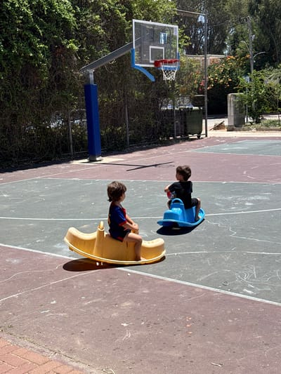 Two children play on playground rockers on a basketball court. One rocker is yellow and the other is blue. The child on the yellow rocker wears a blue and red shirt, while the other child wears black. A basketball hoop stands to the left, partly obscured by greenery. The court surface is cracked and marked with lines. Additional vegetation can be seen in the background.