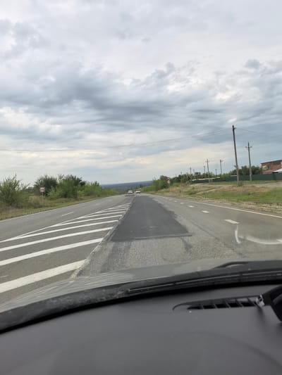 The image shows a view from the interior of a car looking out at a two-lane road in an overcast, rural setting. The road has white lane markings, including chevron markings on the right side. There is a patched section of asphalt in the center of the road ahead. Two white cars are visible in the distance on the road. To the left of the road, there is a sign with a red circle and a horizontal bar, indicating a restriction. Further to the left are green bushes and trees. To the right of the road, there is a grassy area with scattered trees and several utility poles with wires running between them. In the far distance, there appears to be a building with a sign that is difficult to read. The sky is heavily clouded with gray tones. The foreground shows the car's dashboard and windshield wipers. The location is Belogorskiy, Russia.