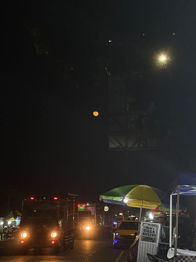 At night in the city of Tanggul, Indonesia, a full moon hangs in the dark sky above a street scene. The moon, colored a warm orange, is positioned to the left of center. The street is illuminated by the headlights of vehicles and the lights of roadside stalls. In the foreground, a truck with its headlights on is visible. To the right, there are two stalls with colorful umbrellas providing shade. One stall has a sign that reads "Resep Cilok Auliya." A person wearing a cap is standing by this stall. Another truck with its lights on is further down the street. The environment appears to be a busy street with food vendors and vehicular traffic.