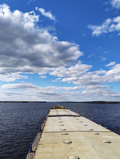 The image shows a barge on a body of water under a blue sky with scattered clouds. The barge is gray and has a flat surface with evenly spaced hatches. In the distance, a shoreline with trees is visible. The location, Bessolovo, Russia, is not visually verifiable in the image.