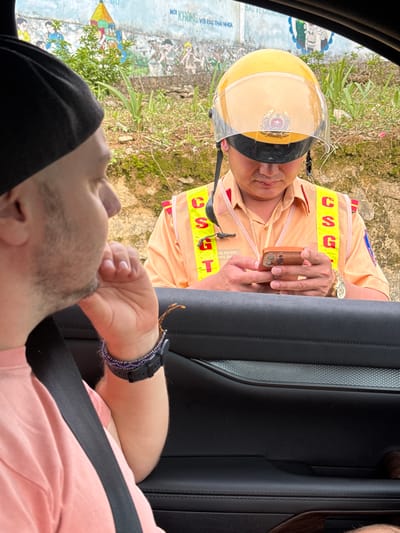 A man wearing a black cap and a pink shirt is seated in a car, wearing a seatbelt. He is looking up at a traffic police officer who is holding a cell phone towards him. The police officer is wearing a yellow helmet with a dark visor, and a yellow traffic vest with red lettering on it. He is also wearing a gold watch. The scene appears to be outdoors, in the city of Quản Bạ Commune, Vietnam, with a wall or sign visible behind the police officer, along with some greenery and dirt. The car door is visible to the side of the driver.