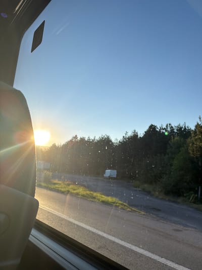 The image shows a view from inside a vehicle, looking out through the windshield. The sun is low on the horizon, casting a bright glare and lens flare across the scene. The sky is a clear, pale blue. On the left side of the frame, a portion of the vehicle's interior, likely a headrest and window frame, is visible.
Outside the vehicle, a paved road runs from the foreground towards a line of dark green coniferous trees. The road has a white line marking its edge. To the left of the road, there is a grassy verge. In the distance, beyond the verge and road, a white van is parked or stopped, partially obscured by the trees. The sun appears to be setting behind the trees, contributing to the warm, golden light illuminating the scene.
The image does not provide any information that would allow for a definitive determination of the location being the city of Gorno Varshilo, Bulgaria.