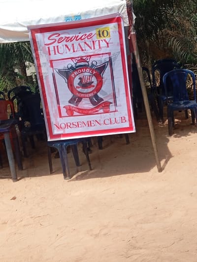An outdoor scene in Okpatu-Ukehe, Nigeria, features a white and red vertical banner prominently displayed, secured under a white tent canopy. The banner reads "SERVICE TO HUMANITY" at the top and "NORSEMEN CLUB" at the bottom, with a central emblem depicting a red circle containing a Viking longship, flanked by crossed axes, and bearing the text "PROUDLY NORSEMEN" and "SERVICE TO HUMANITY." The banner appears slightly creased. Numerous plastic chairs are arranged in the vicinity; several black and one red chair are partially visible on the left, while multiple blue chairs are visible on the right, some grouped together. The ground consists of light brown sand with visible indentations. Green foliage is present in the background, suggesting a vegetated outdoor environment. The scene is illuminated by bright daylight, casting distinct shadows from the chairs and a supporting wooden pole on the right. No individuals are present in the frame.