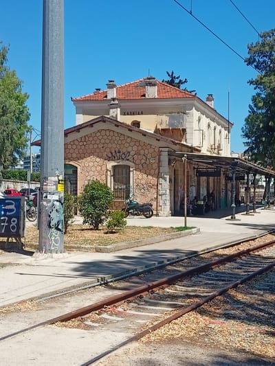 The image depicts the train station of Chalkida in the Municipal Unit of Anthidon, Greece. A train track runs in the foreground, with rusted rails and ballast. To the right of the tracks is a paved platform with a covered waiting area supported by pillars. Several people are seated on a bench under the canopy, and there are signs with numbers on the pillars, possibly indicating platform numbers. The station building itself is a two-story structure with an orange tiled roof. The lower part of the building is clad in stone, while the upper part is painted a pale yellow and exhibits signs of aging. Graffiti is visible on the stone facade and on a utility pole in the foreground. Behind the station, trees and other vegetation are present, and a modern building is visible in the distance. A motorcycle is parked near the station entrance, and a few cars are parked further back. A lamppost with graffiti covers it stands prominently in the left foreground. The sky is clear and blue.